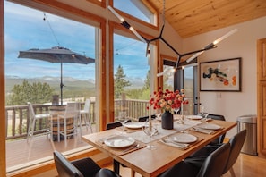 Un comedor con una mesa dispuesta para cuatro personas, un florero con flores y vistas a las montañas a través de grandes ventanales.