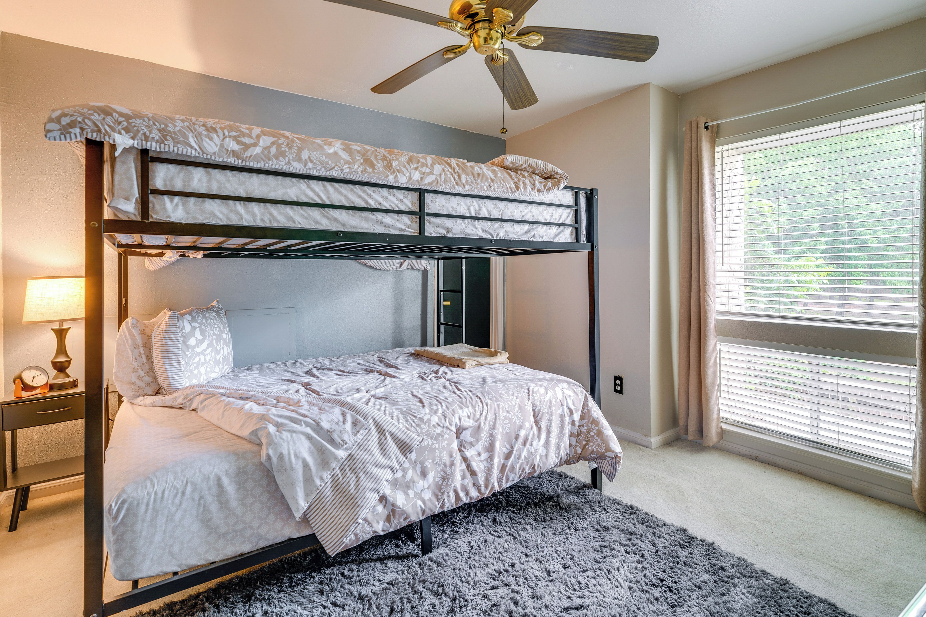 A bunk bed room with a ceiling fan, bedside table with lamp, and a window with blinds.