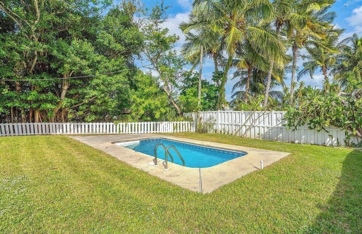 Ein rechteckiger Swimmingpool mit zwei Leitern, umgeben von einem weißen Zierzaun, eingebettet in einen grünen Garten mit Palmen und üppiger Vegetation im Hintergrund.