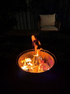 Una xemeneia amb flames, una cadira de vimet i un coixí en un ambient exterior fosc.