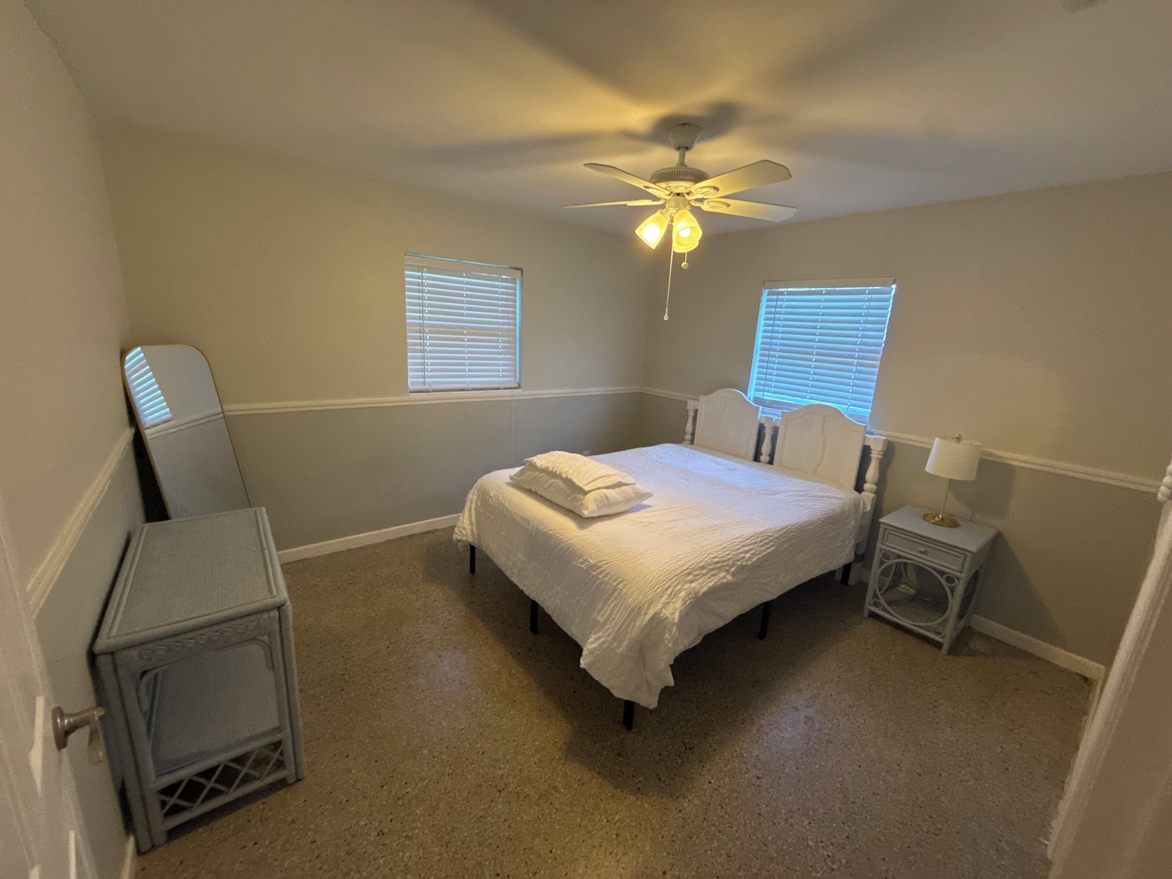 A bedroom with a bed, a ceiling fan, two windows with blinds, a nightstand, and a dresser.