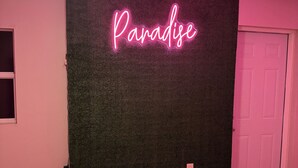 Huoneessa on kattotuuletin, neonvalomainos, jossa lukee ”Paratiisi”, ovi ja laattalattia.