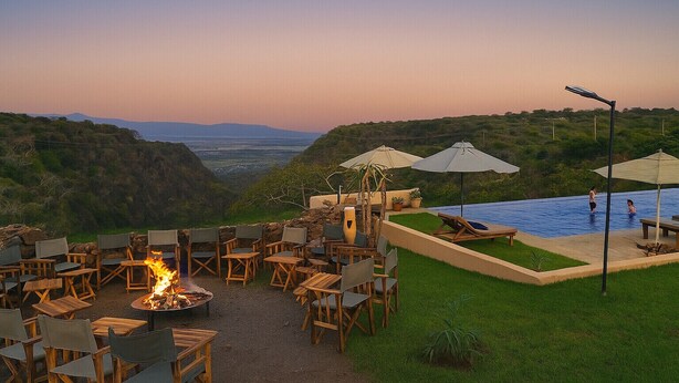 Un espacio al aire libre con sillas, mesas, una chimenea y una piscina con sombrillas.
