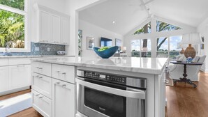 Een moderne keuken met witte kasten, een groot eiland met een blauwe kom, een ingebouwde oven en uitzicht op het buiten.