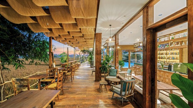 Un restaurante con mesas y sillas de madera, una zona de bar con estanterías llenas de botellas y amplias ventanas que dan a un campo.