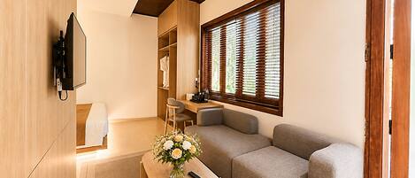 A modern living room with a sofa, a wooden coffee table, a TV mounted on the wall, and a window with blinds.