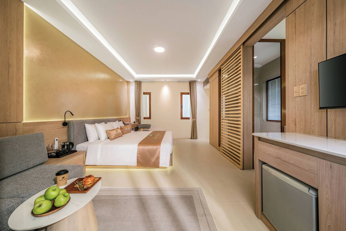 A modern hotel room with a bed, a sofa, a dining table with apples, a coffee cup, and a TV.
