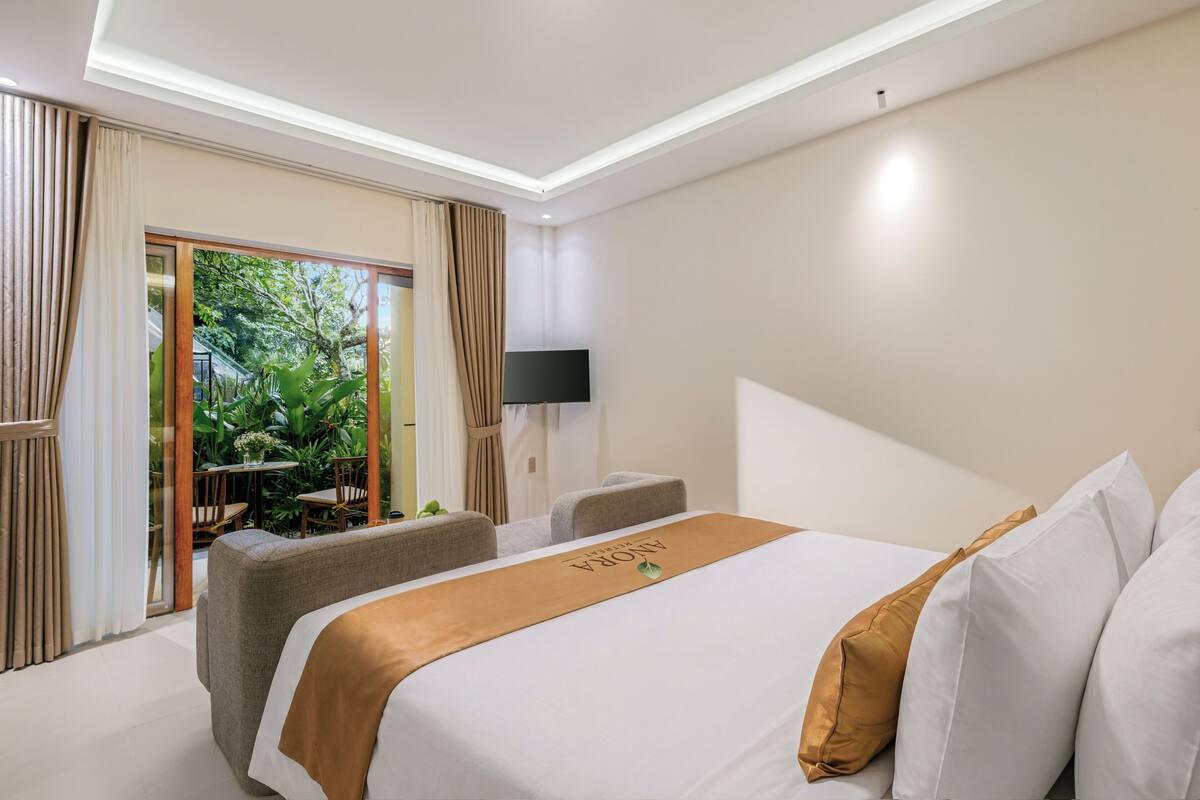 A hotel room with a bed, a TV, a balcony with plants, and a view of greenery.