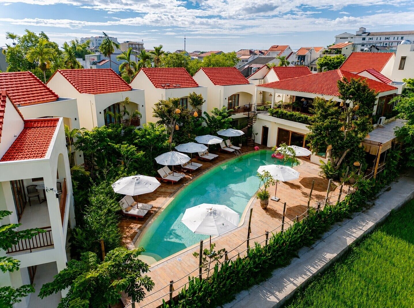 A hotel with a swimming pool surrounded by lounge chairs and umbrellas, with red-tiled roofs and lush greenery.