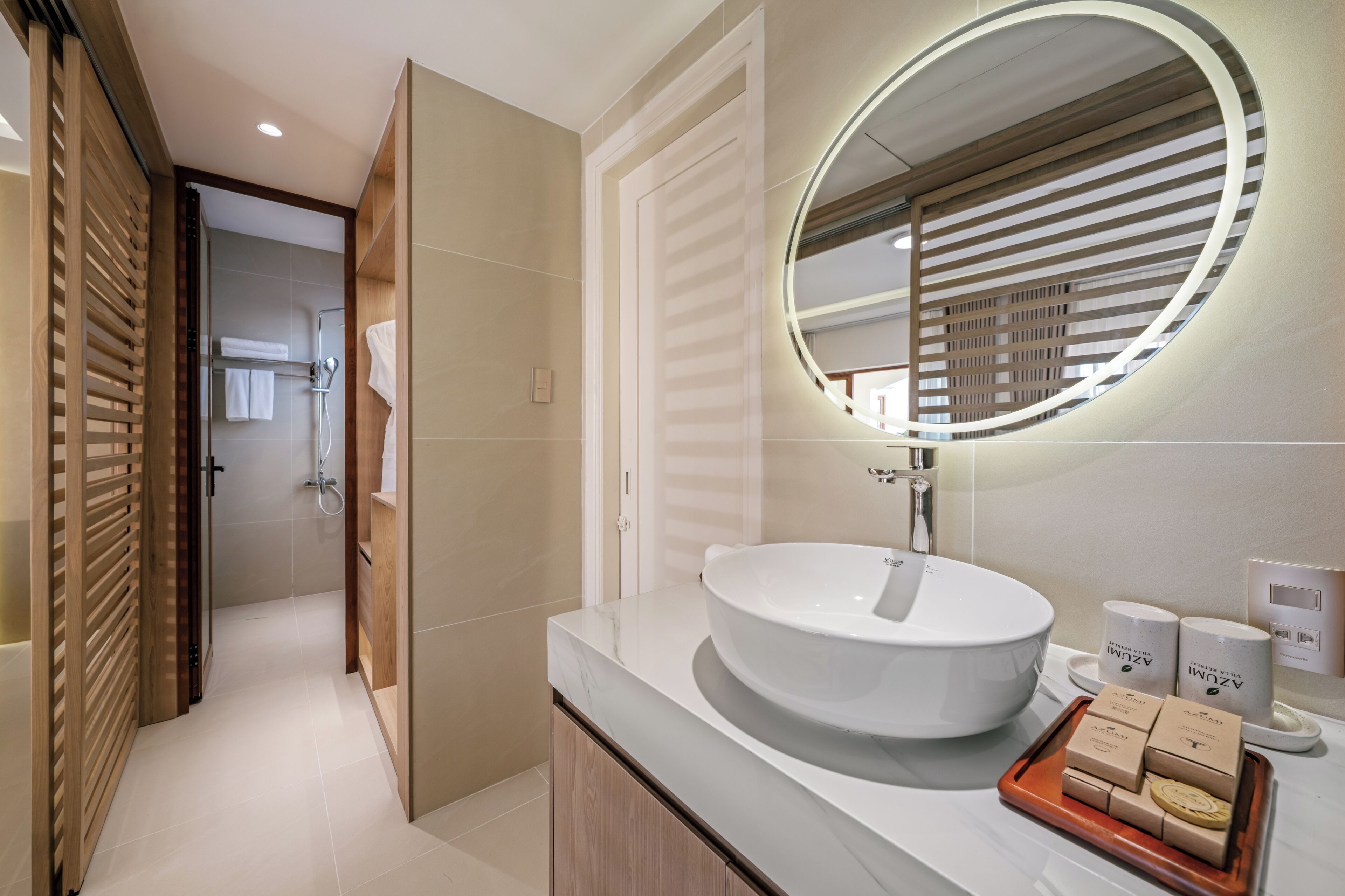 A modern bathroom with a large round sink, a mirror with a circular light, and a wooden door leading to a shower area.