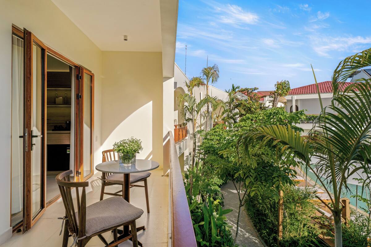 A balcony with a table and chairs, a sliding glass door, and a view of a courtyard with plants and a building in the background.