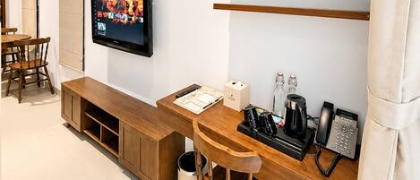 Una habitación de hotel con un escritorio de madera, un televisor montado en la pared, un teléfono, una cafetera y un cubo de basura.