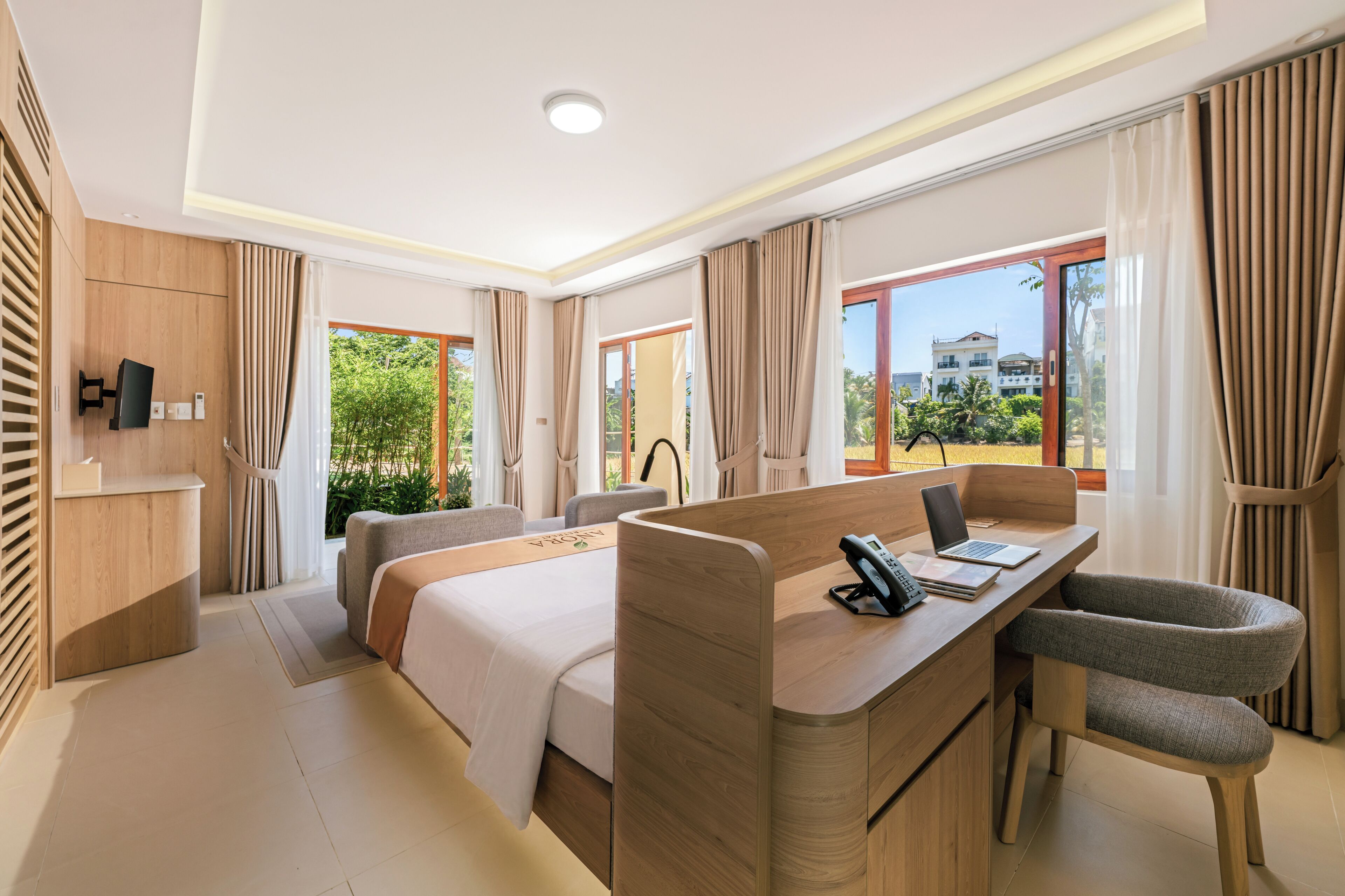A hotel room with a bed, desk, chair, and a view of greenery and buildings.