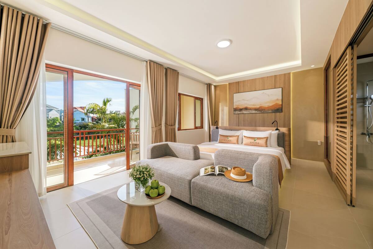 A modern hotel room with a balcony, a bed with pillows, a sofa, a small table with fruits, and a painting on the wall.