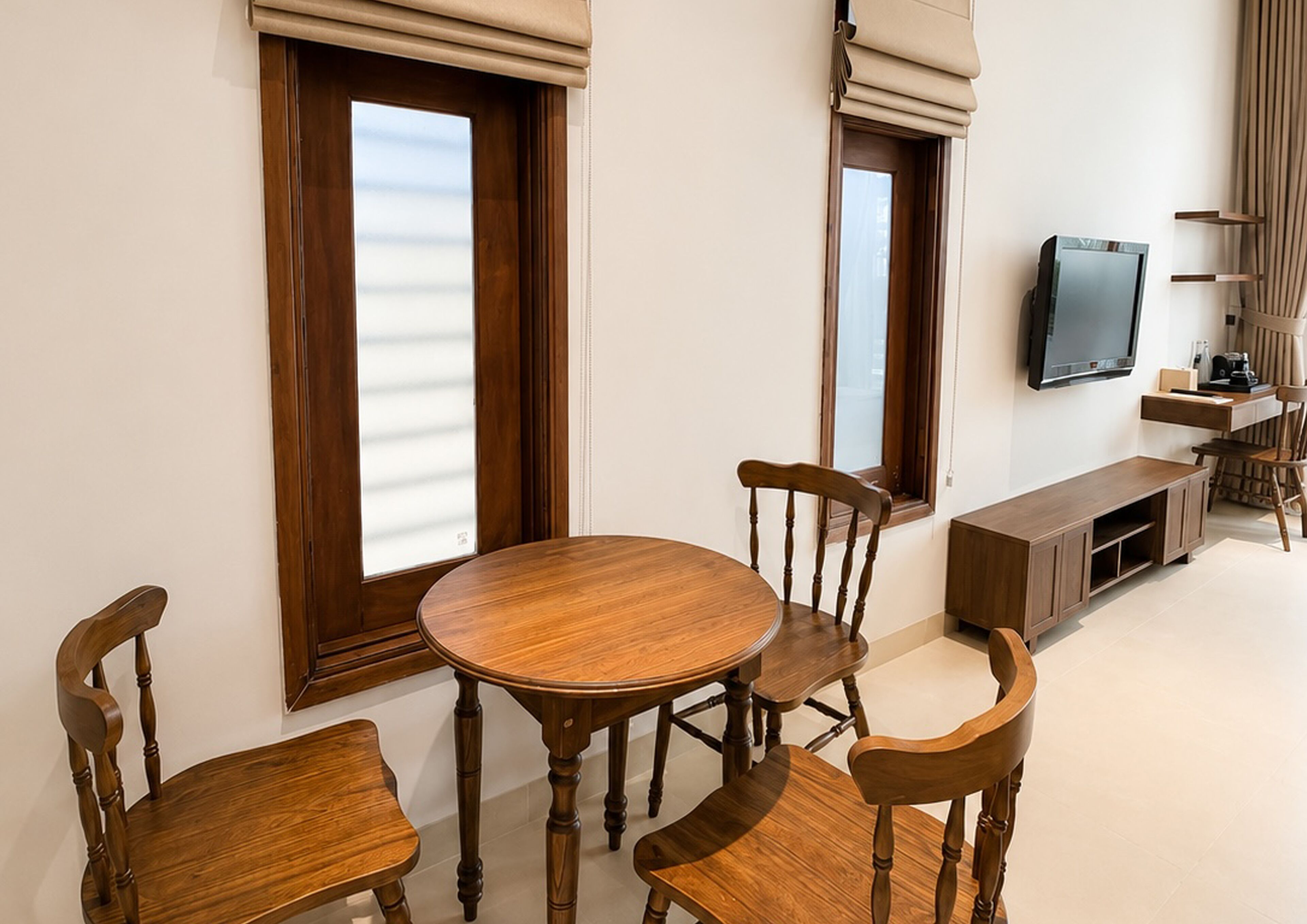 A dining area with a round wooden table and four chairs, a TV mounted on the wall, and a small cabinet with a coffee machine.