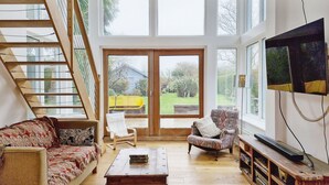 A spacious living area with a wooden staircase, a sofa, a coffee table, a television, and a large window offering a view of the outdoors. - Sleeps 8 guests  in 4 bedrooms (Emsworth)