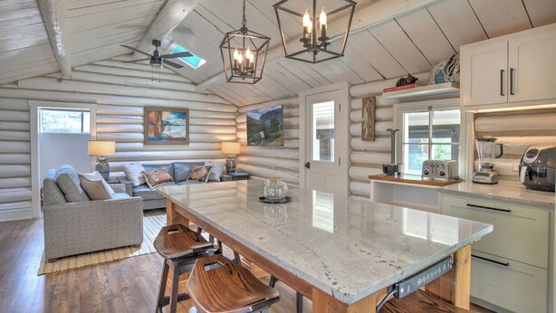 L’intérieur d’une cabane en rondins présente un îlot central, un coin salon, une cuisine et des poutres en bois au plafond.