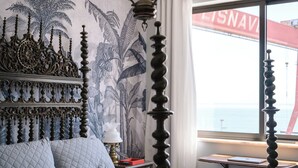 Ein Bett mit Kopfteil, ein Nachttisch mit Lampe, ein Fenster mit Meerblick und eine Wand mit einem tropischen Wandbild.
