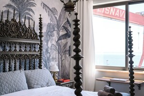A bed with a headboard, a bedside table with a lamp, a window with a view of the sea, and a wall with a tropical mural. - Casa Rio • Wide Estuary Views • Walk To Lisbon Ferry • Free Parking (Almada)