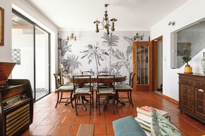 Comedor con una mesa redonda, sillas, una araña y una pared de azulejos con motivos de palmeras.