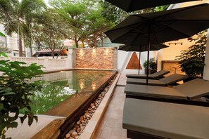 Una piscina con sillones de descanso y una sombrilla, rodeada de árboles y un muro de piedra.