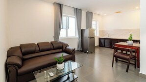 Una sala de estar moderna con un sofá marrón, una mesa de centro de vidrio, un comedor con una mesa y sillas de madera, una cocina con refrigerador y una ventana con cortinas.
