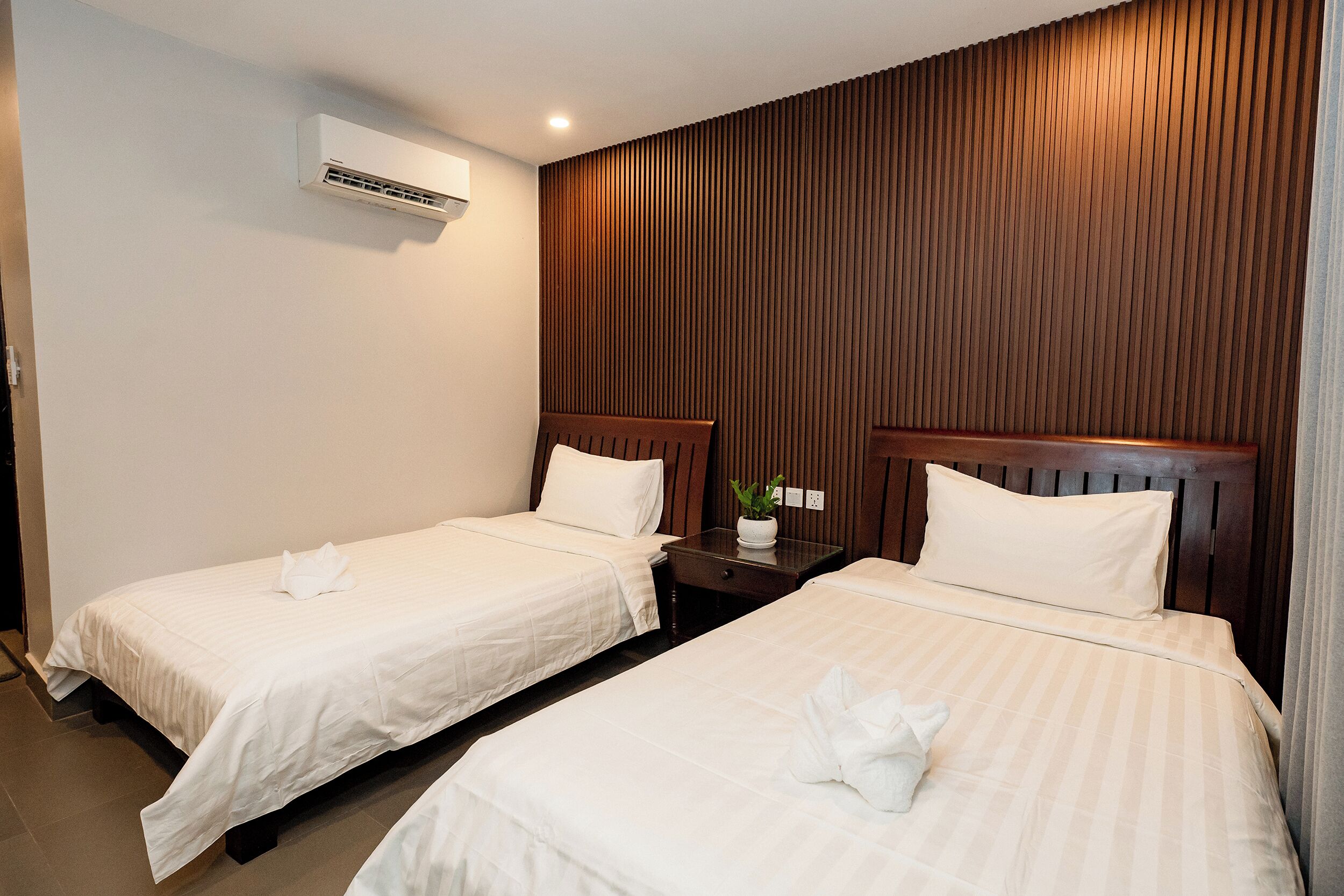 Quarto de hotel com duas camas, mesa de cabeceira, planta em vaso e ar condicionado.