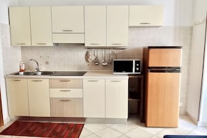 Una cocina con armarios de tonos claros, microondas, refrigerador, lavamanos y una alfombra roja.