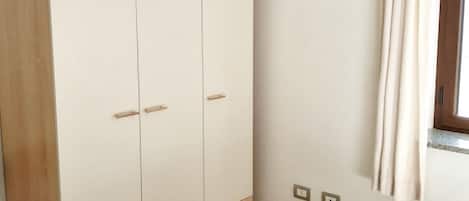 A bedroom with a wardrobe, a bed, a window with curtains, and a wall-mounted light switch.
