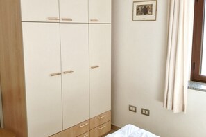 A bedroom with a wardrobe, a bed, a window with curtains, and a wall-mounted light switch. - Cala Gonone, 4-room apartment (4th floor) (Cala Gonone)