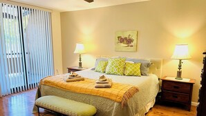 Un dormitorio con una cama, dos mesitas de noche, un banco, un ventilador de techo y una ventana con persianas.