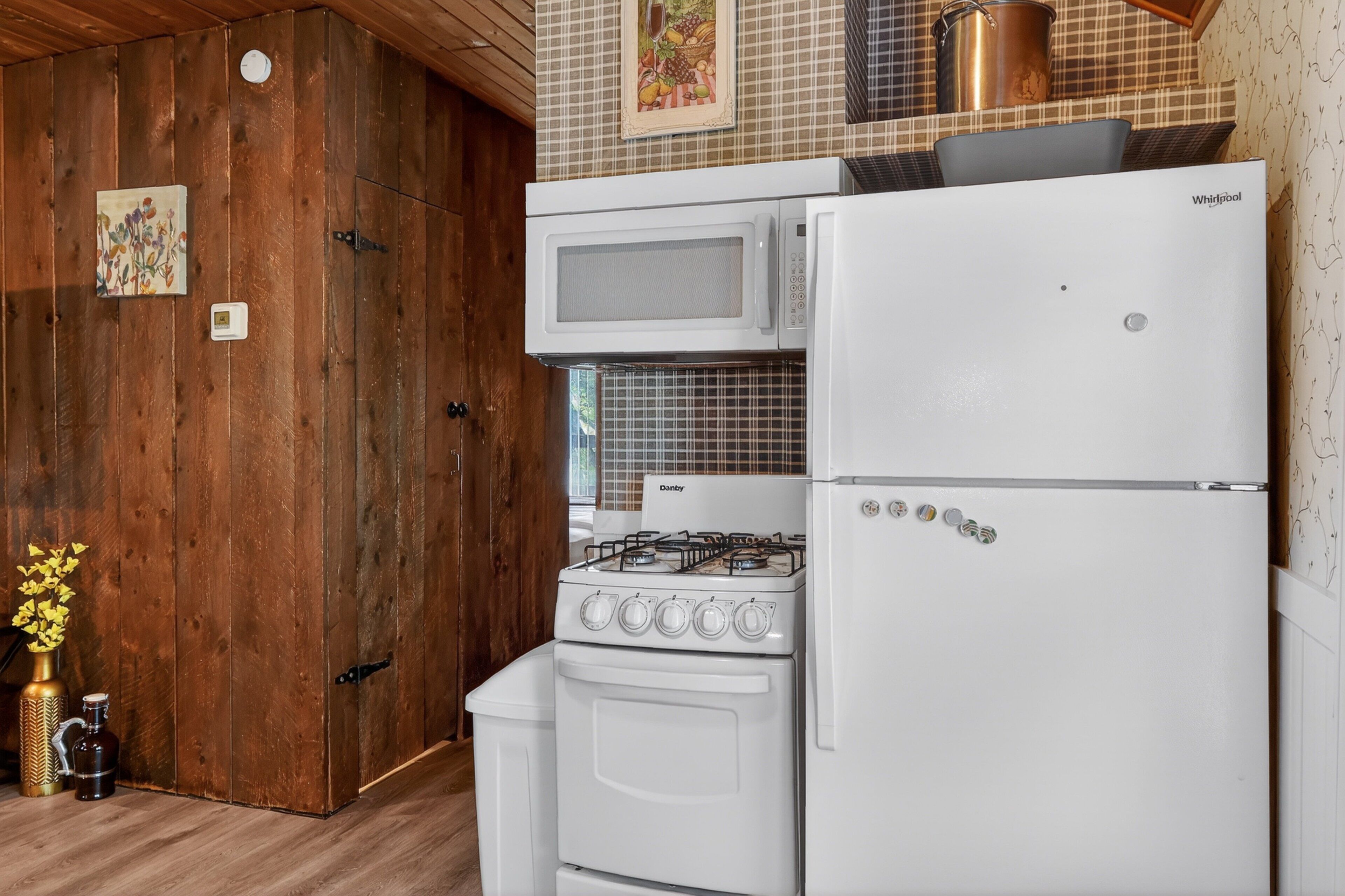 A kitchen with a stove, microwave, and refrigerator, wooden walls, and a small vase with flowers.