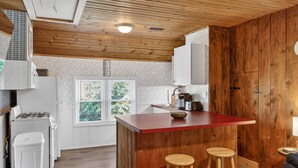 A kitchen with wooden ceiling, white cabinets, a red island, two stools, and a window with a view of greenery. - City Explorer Basecamp Buffalo 1 Bedroom Duplex with AC (Buffalo)