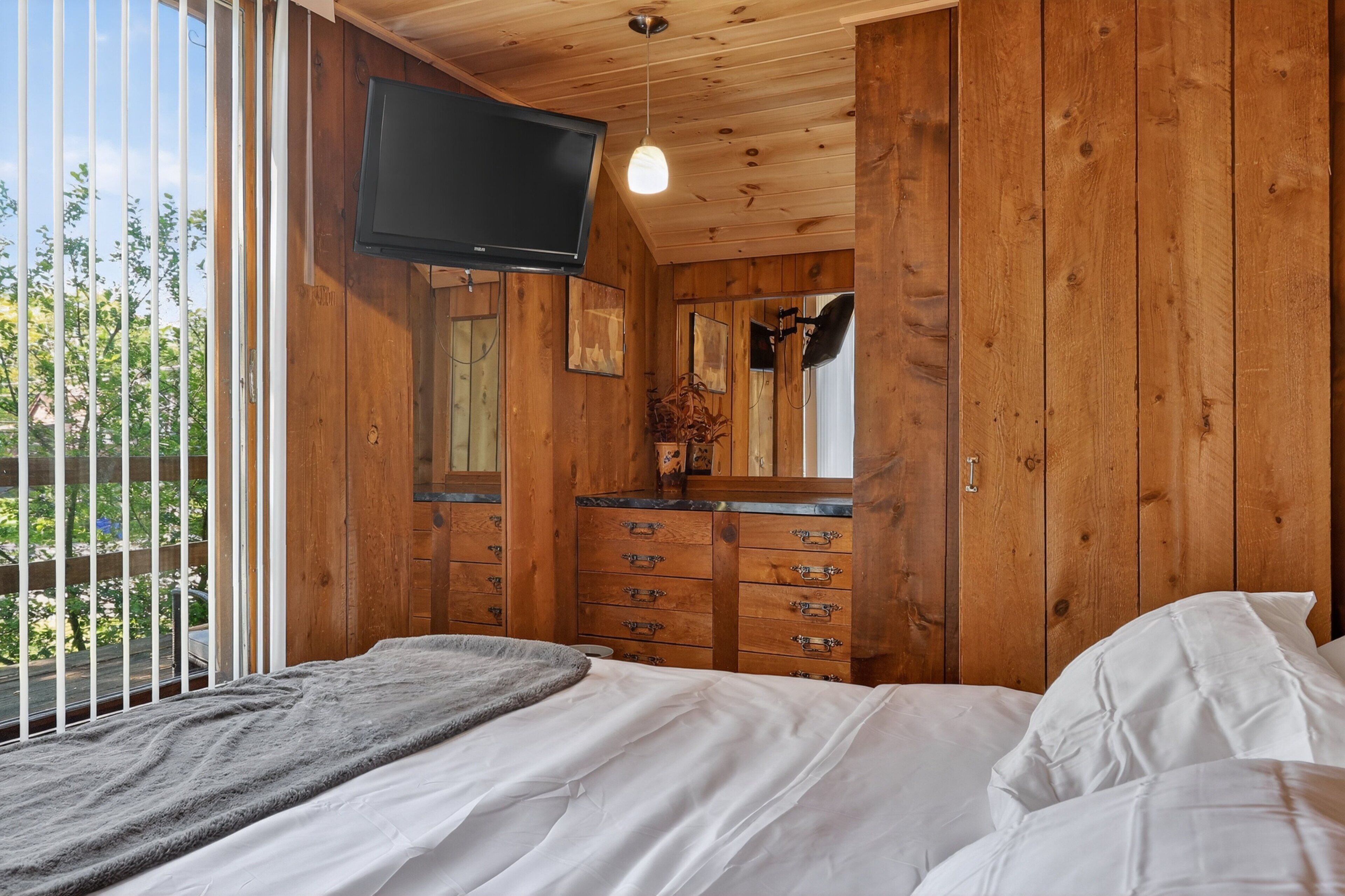 A wooden-paneled bedroom with a bed, a dresser, a television, and a window with a view of greenery.