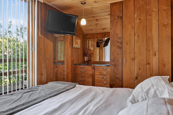 Ein Schlafzimmer mit Holzwandverkleidung, einem Bett, einer Kommode, einem Fernseher und einem Fenster mit Blick auf Grünflächen.