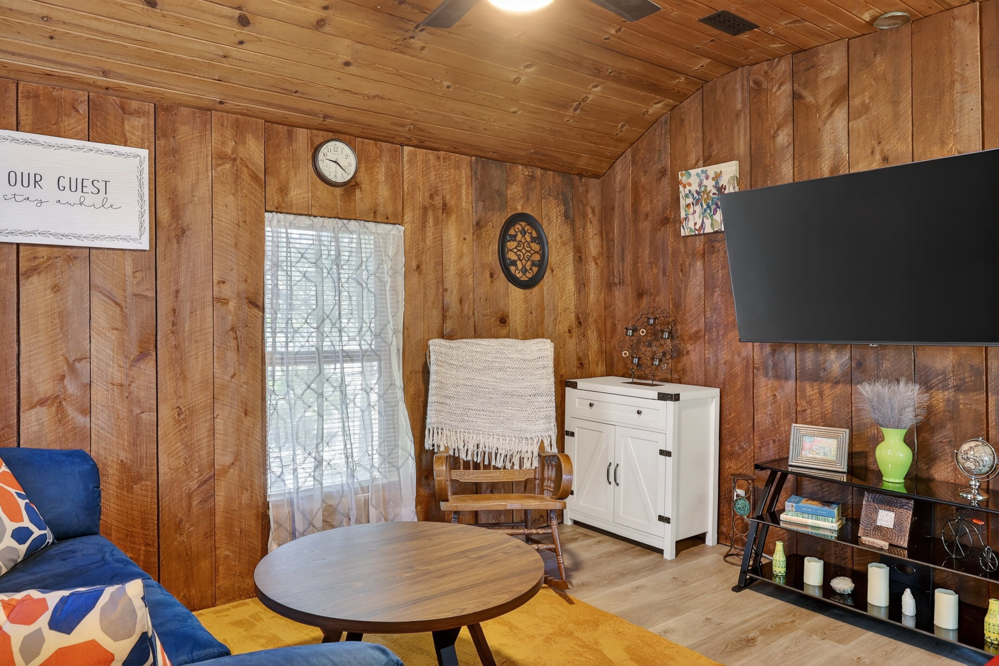 A wooden-paneled room with a round table, a blue sofa, a white cabinet, and a clock on the wall.