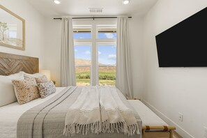 A bedroom with a large bed, a TV mounted on the wall, and a view of a landscape through the window. - Grand Retreat At Coral Canyon 7 Bedroom Home (Washington)