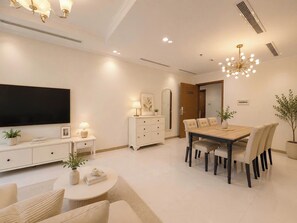 Een moderne woonkamer met een flatscreen-televisie, een eethoek met een houten tafel en stoelen, een witte console en een kroonluchter.