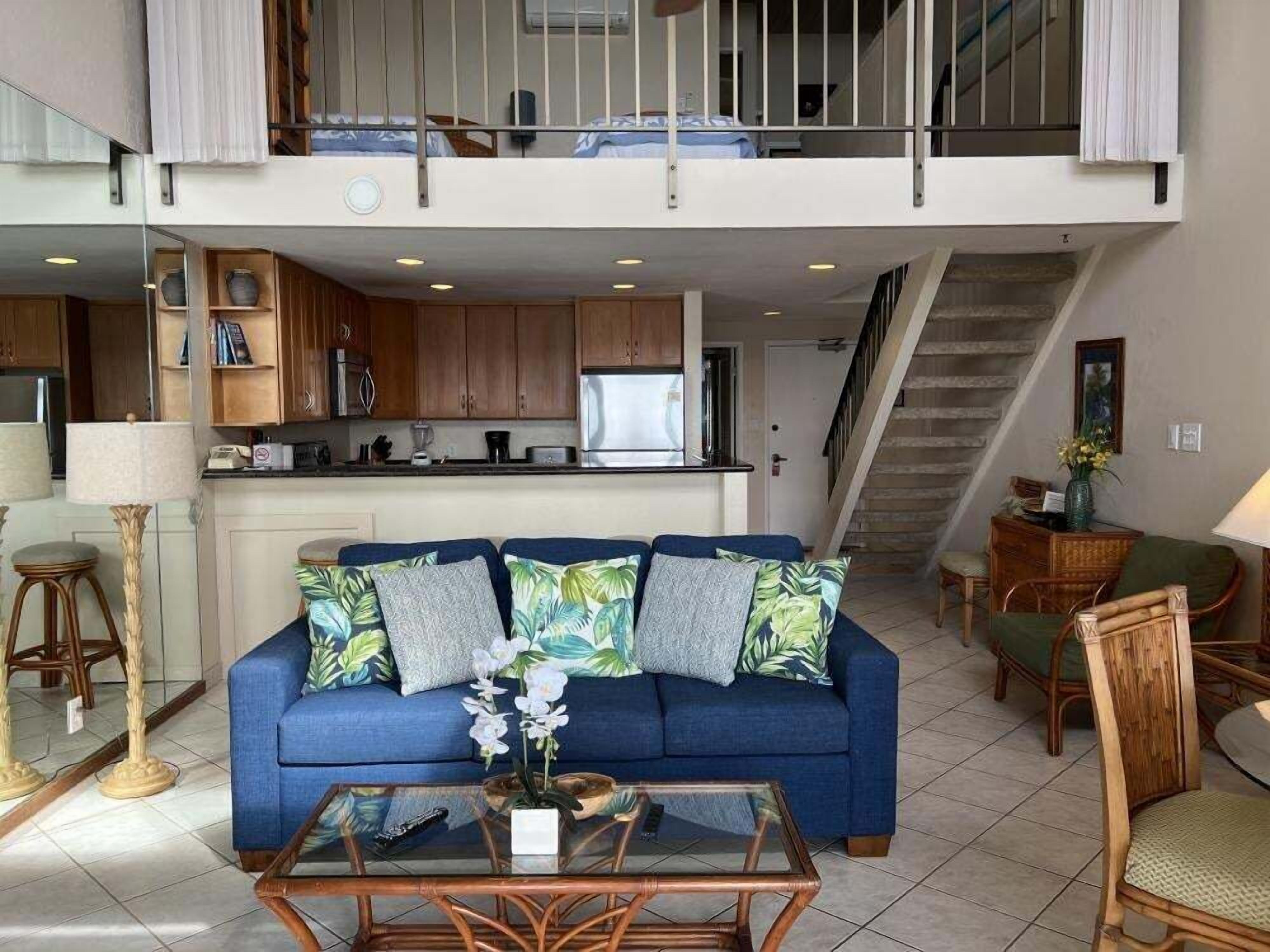 A living room with a sofa, coffee table, and chairs, a kitchen in the background, and a staircase leading to an upper level.