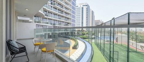 A balcony with a view of a sports field, a seating area with chairs, and a high-rise building in the background.