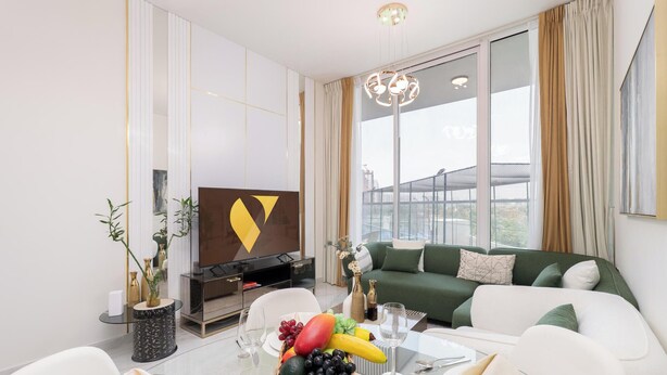 A modern living room with a large window, a green sofa, a flat-screen TV, and a dining area with fruit and glassware.