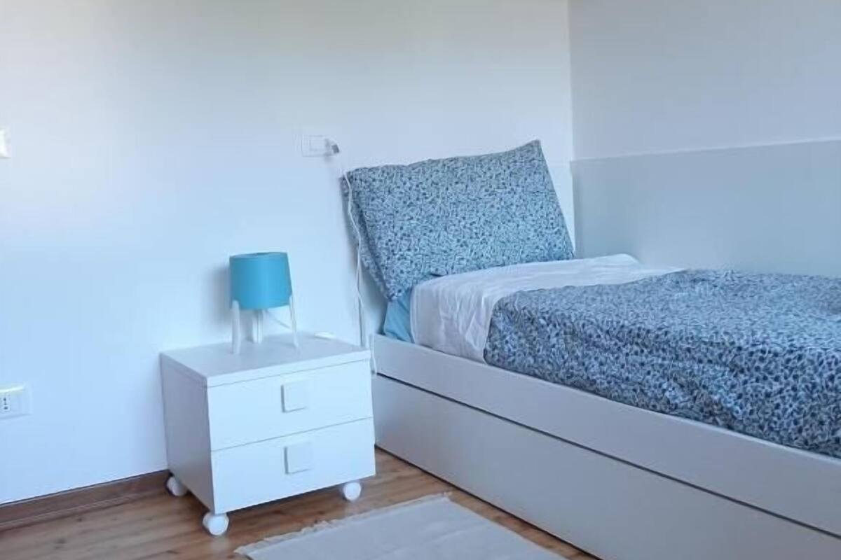 Una camera da letto con un letto, un comodino, un tavolino, un tappeto bianco e un quadro appeso al muro.