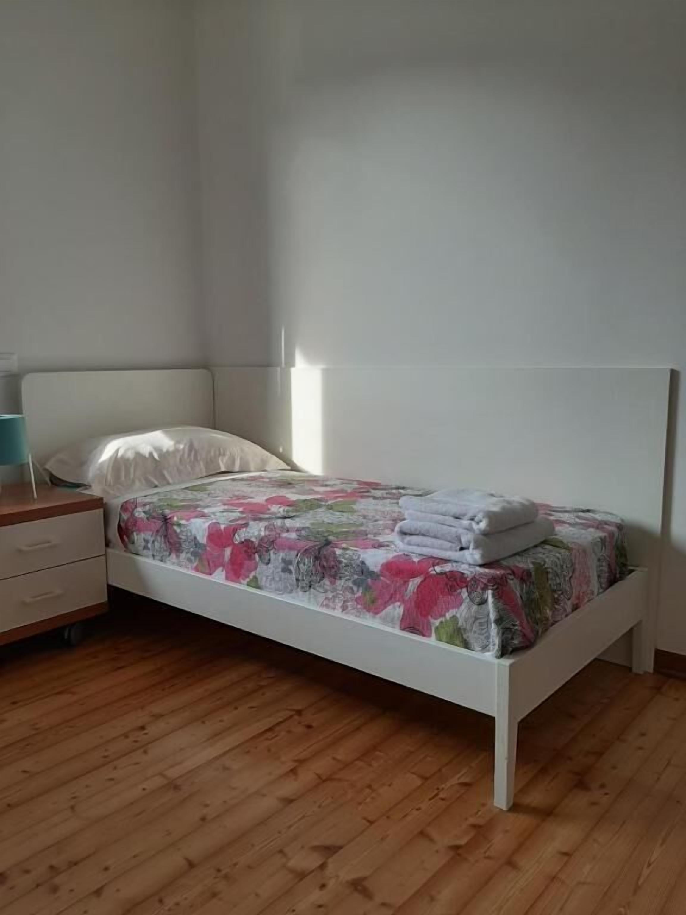 Un lit simple avec une couvre-lit à motifs floraux, une table de chevet avec une lampe et un plancher en bois.