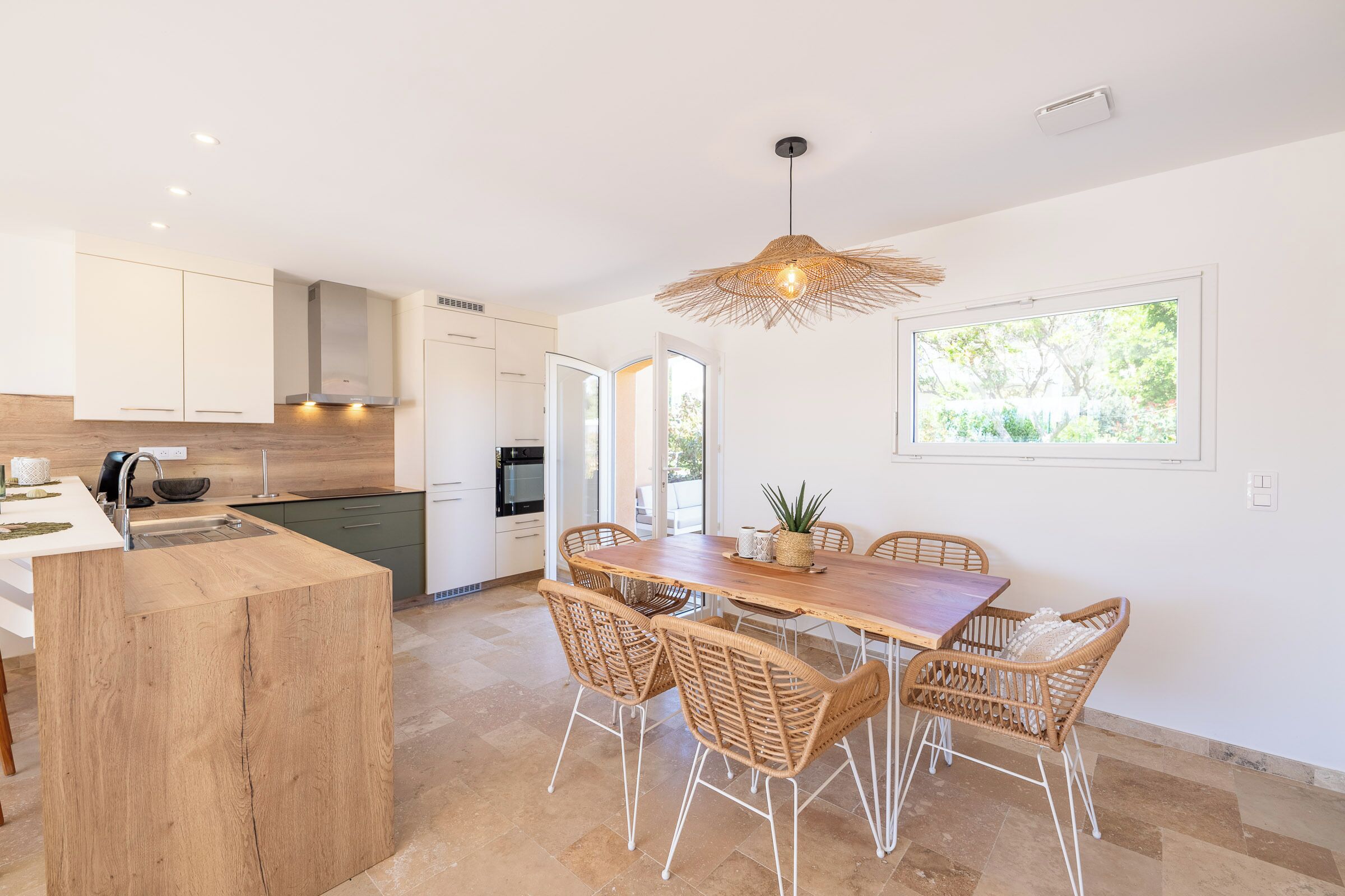 Une cuisine moderne avec des meubles blancs, un îlot central en bois et un coin repas comprenant des chaises en osier et une table en bois.