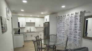 A modern kitchen with white cabinets, a stainless steel refrigerator, and a dining area with a glass table and metal chairs. - Cozy 1-BR/BA cottage in beautiful Stonecrest near I-20 & I-285, Arabia Mountain (Stonecrest)