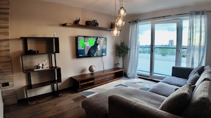 Ein modernes Wohnzimmer mit Flachbildfernseher, Holzregalen, einer Couch und Blick durch ein großes Fenster.