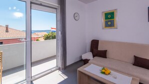 A room with a sofa, a small table with lemons, a clock, and a window with a view of buildings and a flag. - Peaceful Apartments Harfa- One Bedroom Apartment With Balcony and Partial Sea View 2 (Orebic)