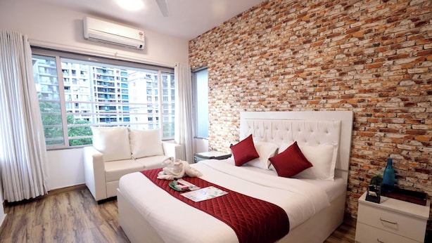 Habitación de hotel con pared de ladrillo, cama grande con almohadas rojas, cabecero blanco, mesita de noche y ventana con cortinas.