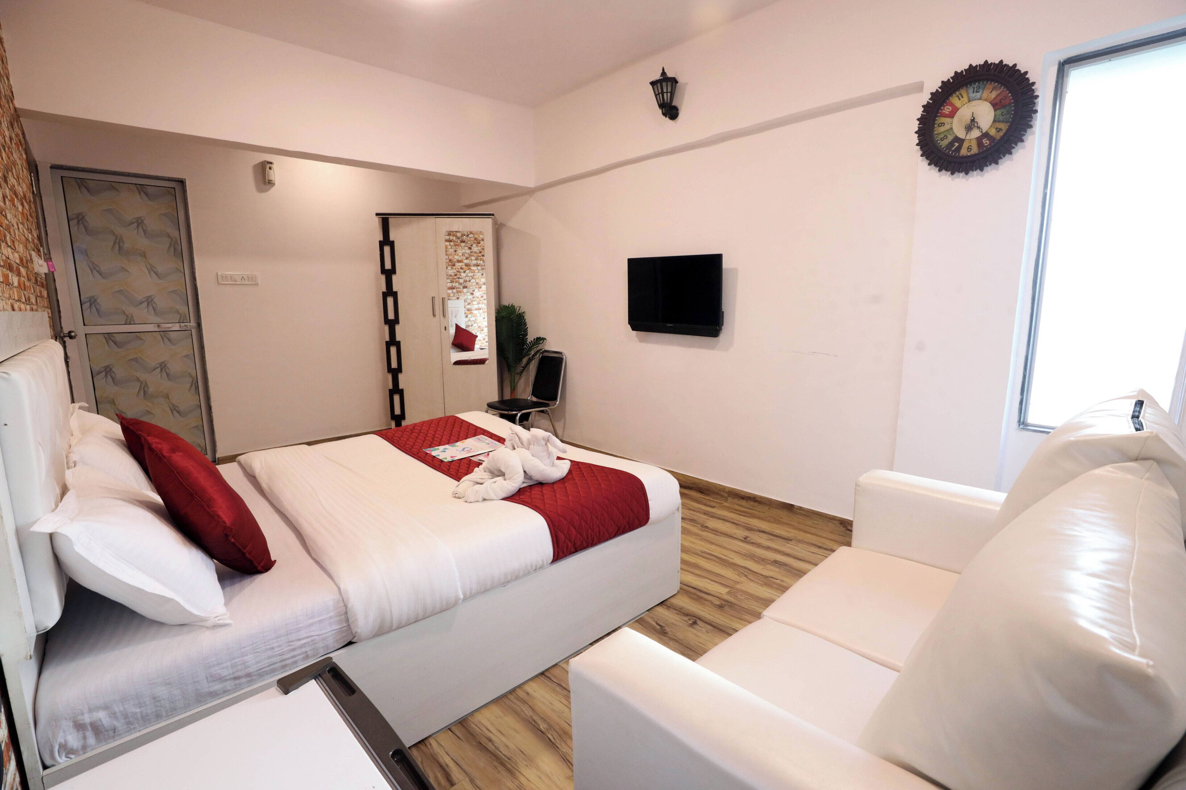A hotel room with a bed, a white sofa, a TV, a clock, and a window.