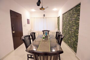 Uma área de jantar com mesa de vidro, cadeiras pretas, ventoinha no teto e parede com textura semelhante a musgo.